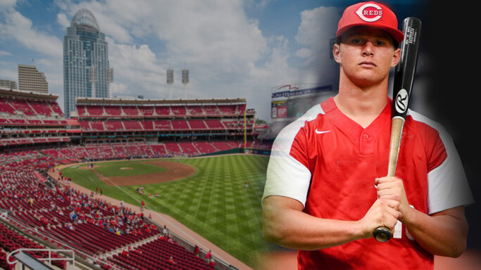 A Cincinnati Reds baseball player holding a bat in front of a stadium.