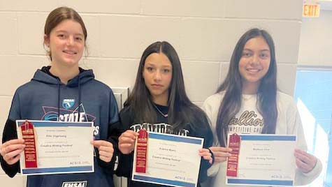 Three girls holding ACSI certificates in front of a wall, celebrating their success in the Creative Writing Contest with awards.