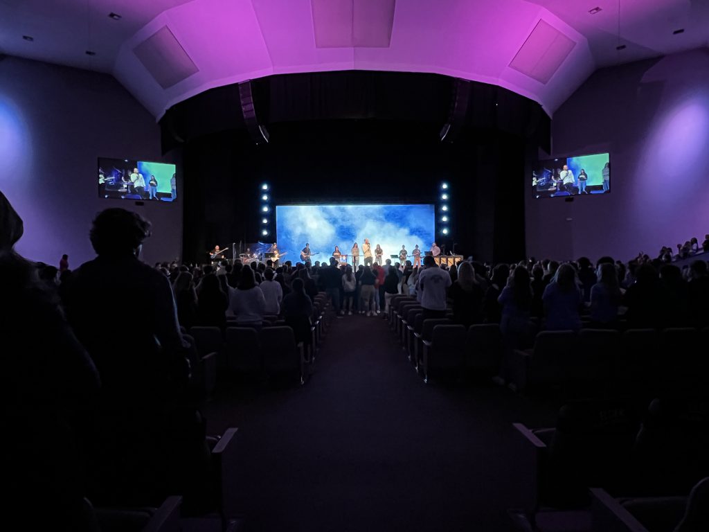 A spiritual church auditorium filled with people watching a large screen with emphasis on the week.