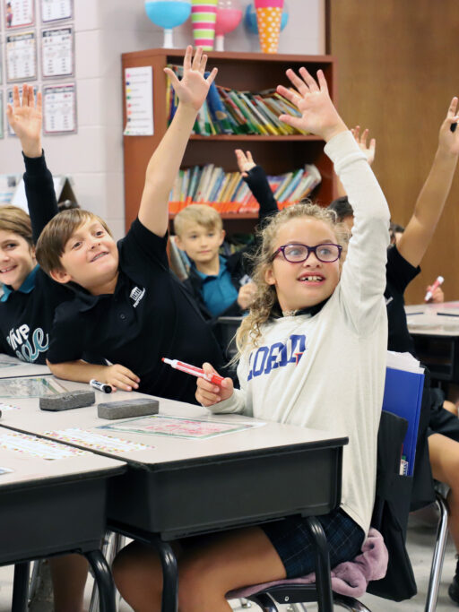 A group of children raising their hands in a classroom, showcasing their enthusiasm for learning and potential for admissions.
