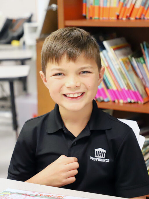 A boy in a black shirt and uniform smiles in front of a bookshelf.