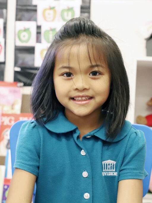A young girl in a blue shirt, wearing a uniform, smiling at the camera.