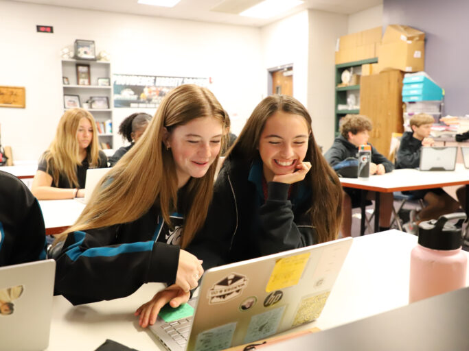 A group of girls utilizing technology, sitting at a desk with laptops in front of them.