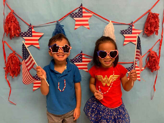 Two children posing in front of an American flag backdrop during a patriotic event.