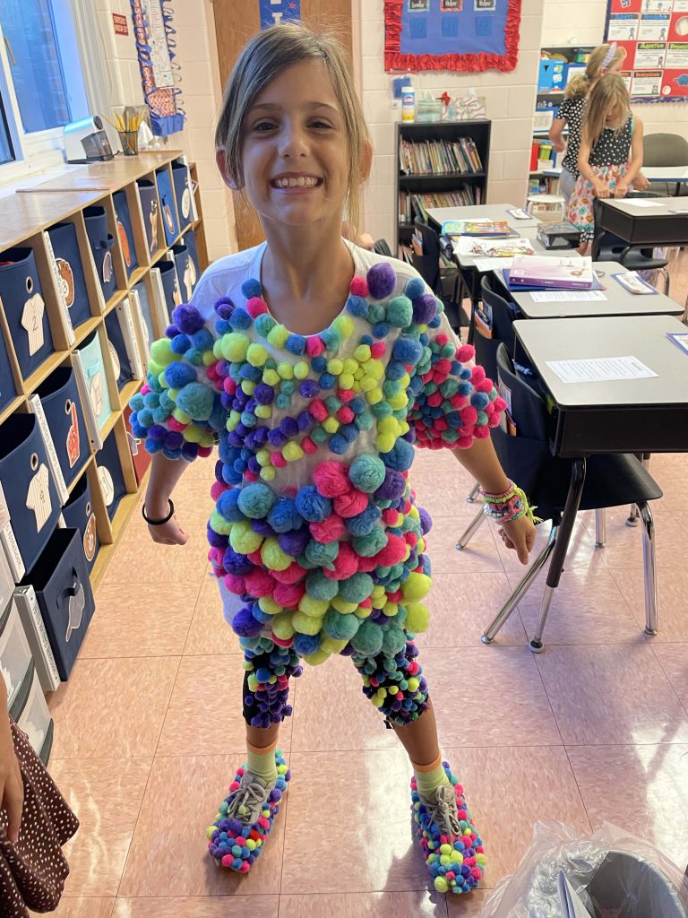 A girl wearing a colorful pom pom costume among students in an international classroom, celebrating.