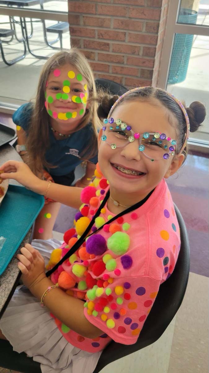 Two students celebrate International Dot Day with colorful face paint, sitting at a table.