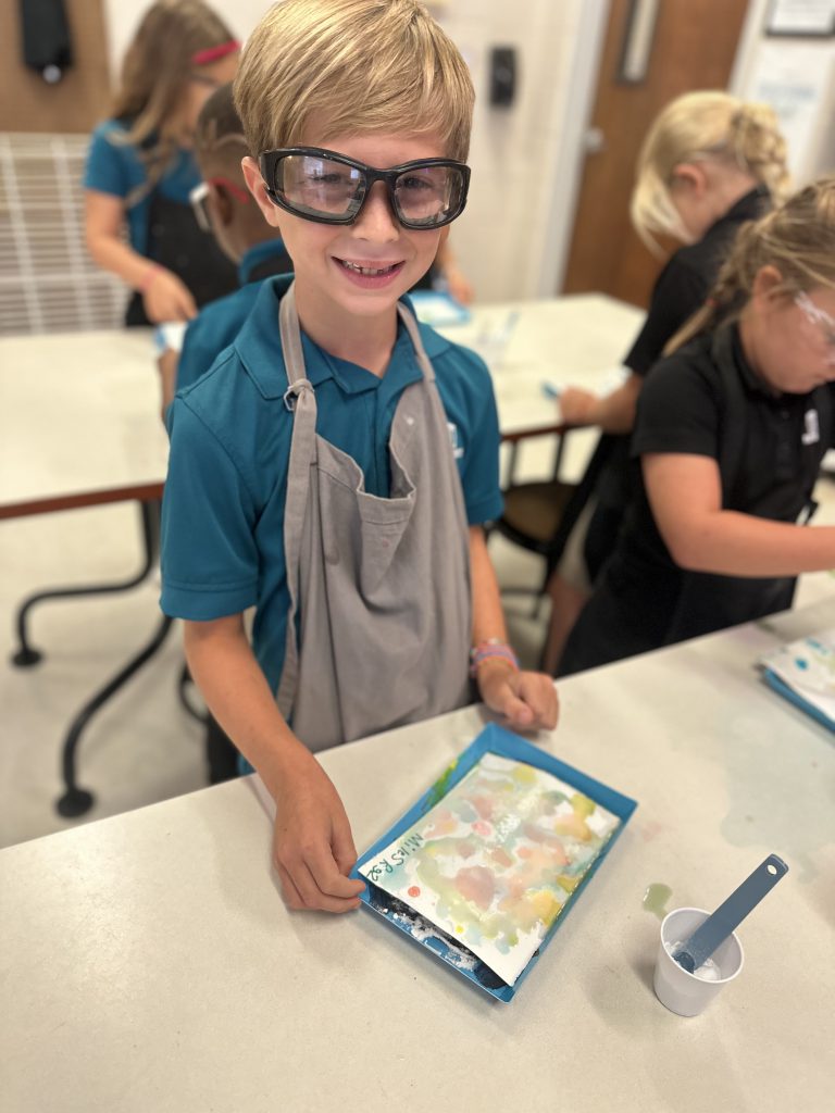 In a Lower School classroom, a young boy wearing glasses and an apron enthusiastically engages in a STEAM Discovery activity.
