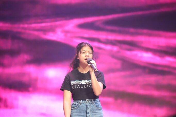 Demi Tebow, a girl in a black shirt, singing into a microphone on the chapel stage.