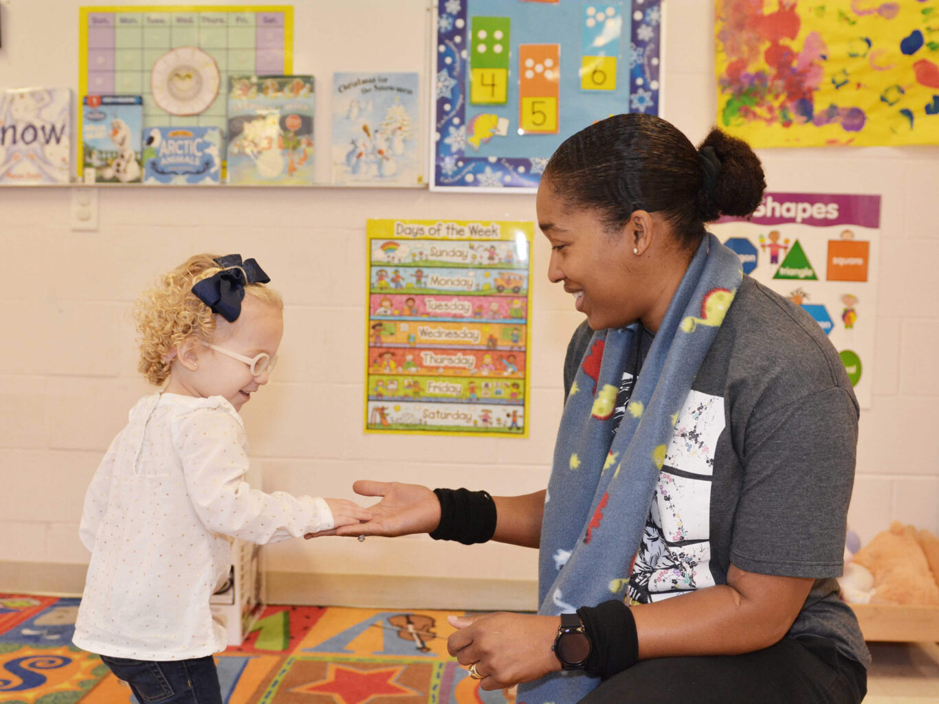 A woman is holding a little girl's hand in a preschool classroom.