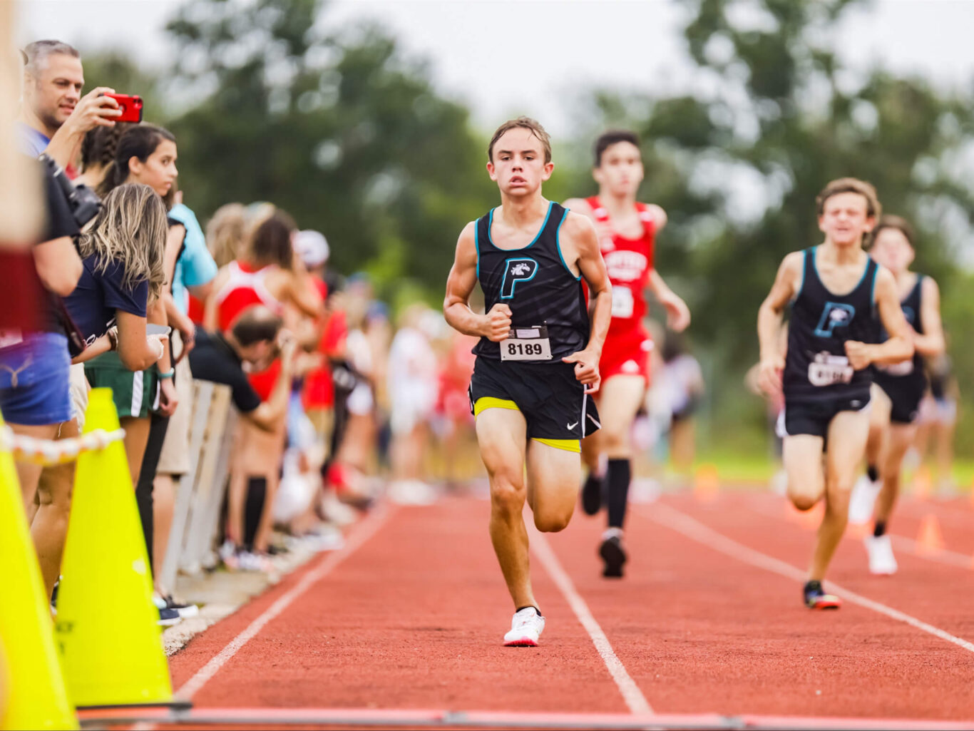 A group of boys running in a cross country race, with spectators cheering them on.