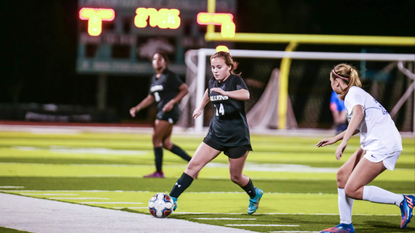 Two girls soccer players kicking the ball on a field.