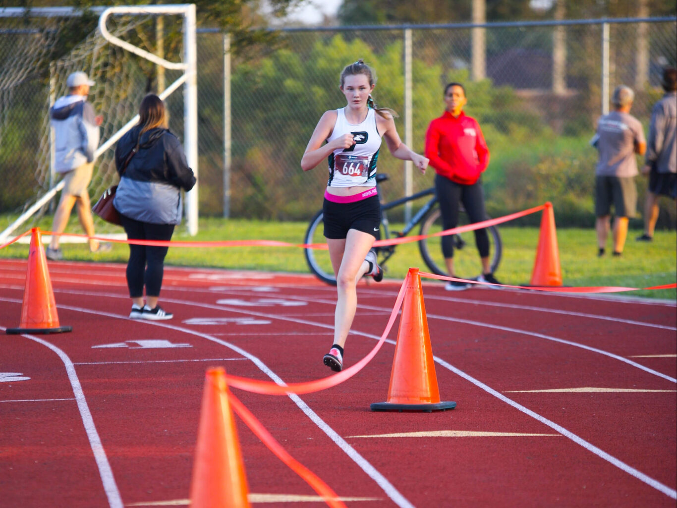 A Cross Country girl is running on a track in front of orange cones.