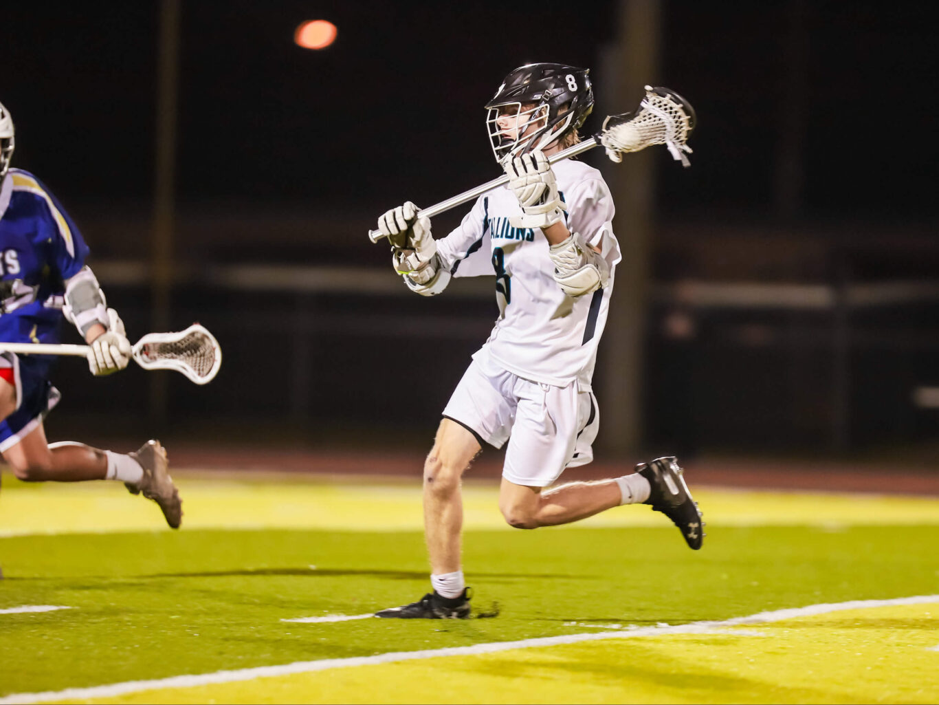 A boys' lacrosse player is running on the field at night.