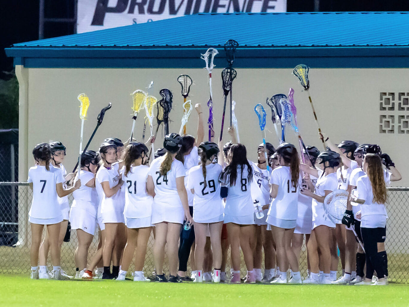 A gathering of girls exuberantly hoisting their lacrosse sticks high in the air.