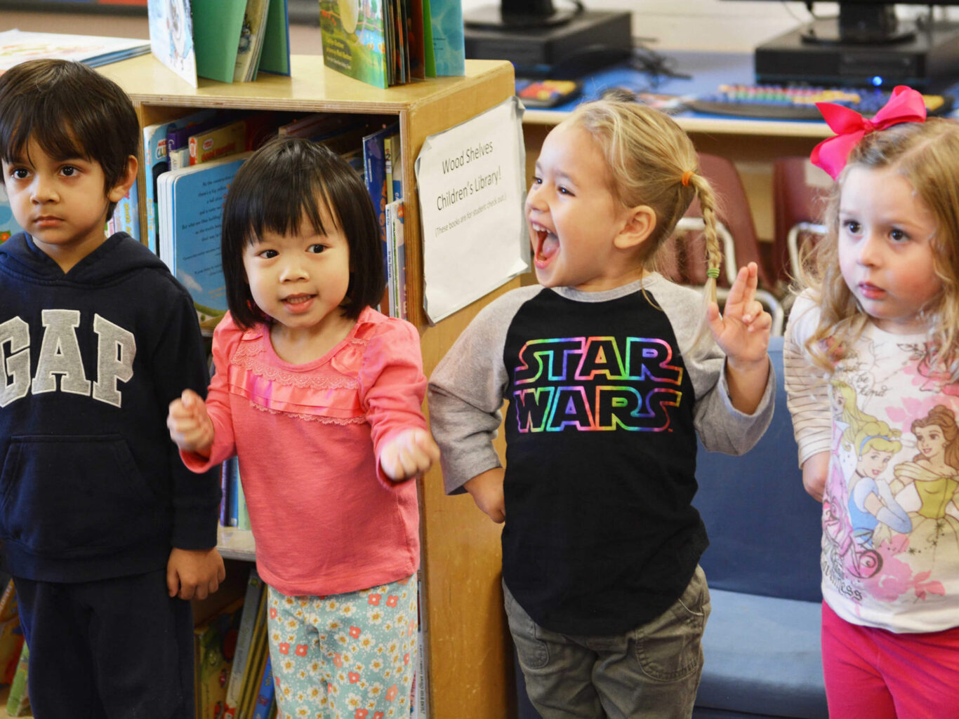 A group of preschool children standing in front of a bookcase.