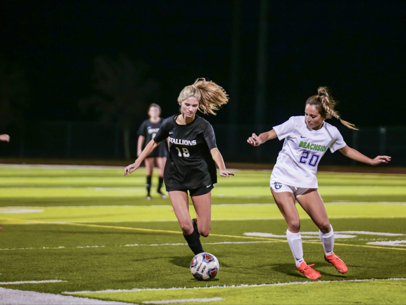 Two girls engaging in a soccer match under the moonlight on a field.