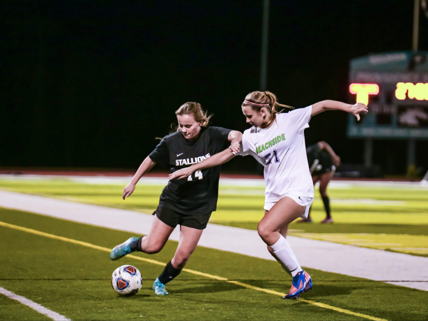 Two girls playing soccer on a field at night.