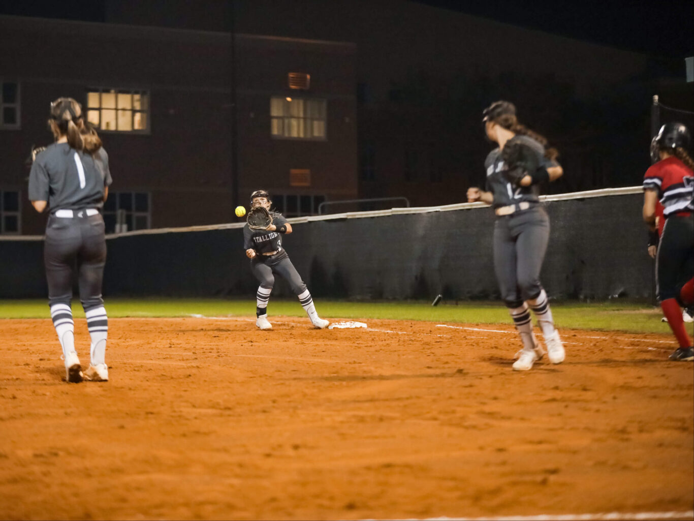 A group of softball girls playing on a field at night.