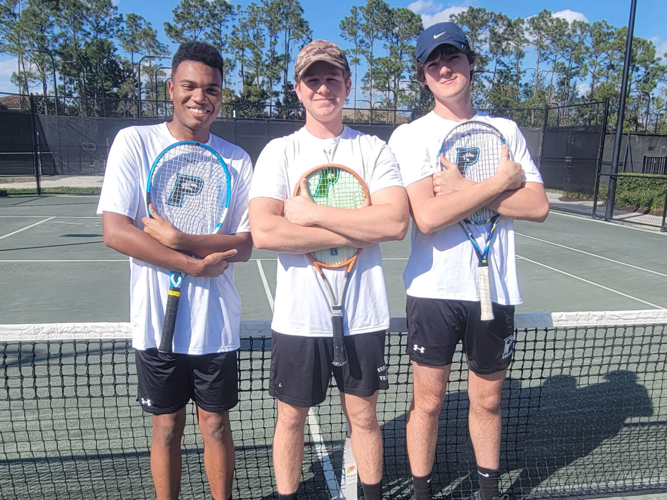 Three boys standing on a tennis court holding tennis rackets.