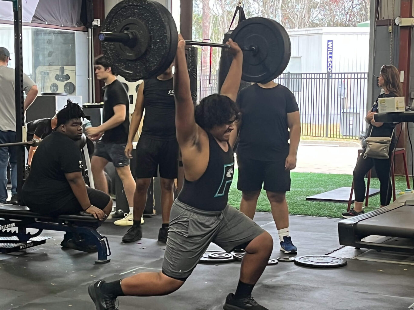 A man weightlifting with a barbell in front of a group of boys.