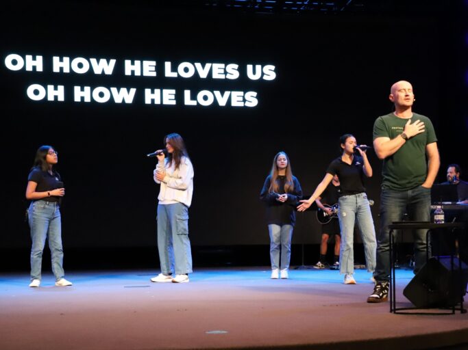 A group of people passionately singing on stage, showcasing their love for humanity through the heartfelt words of "how he loves us.